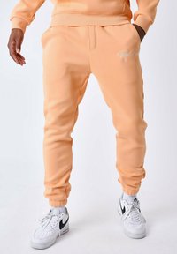 Joggers color durazno hechos de tela suave con puños elásticos, que cuentan con bolsillos laterales y un logo blanco en el muslo izquierdo. Combinados con zapatillas blancas.