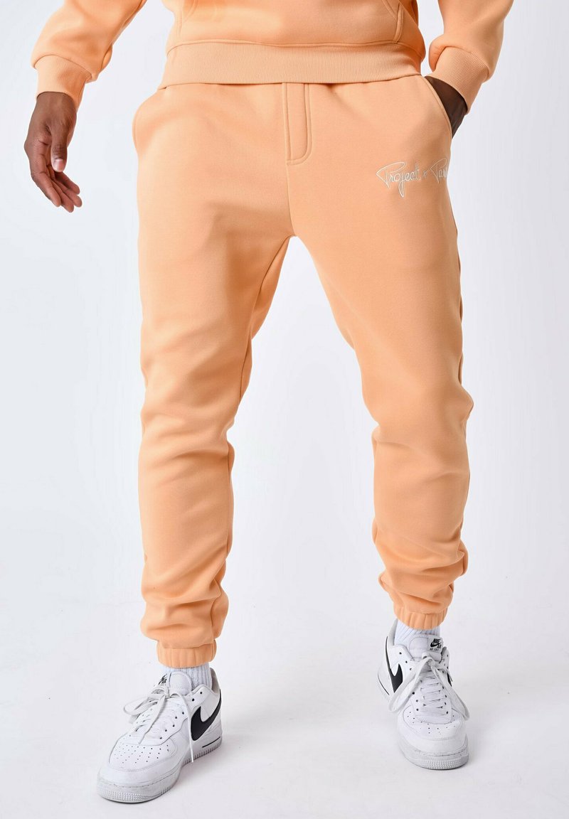 Joggers color durazno hechos de tela suave con puños elásticos, que cuentan con bolsillos laterales y un logo blanco en el muslo izquierdo. Combinados con zapatillas blancas.