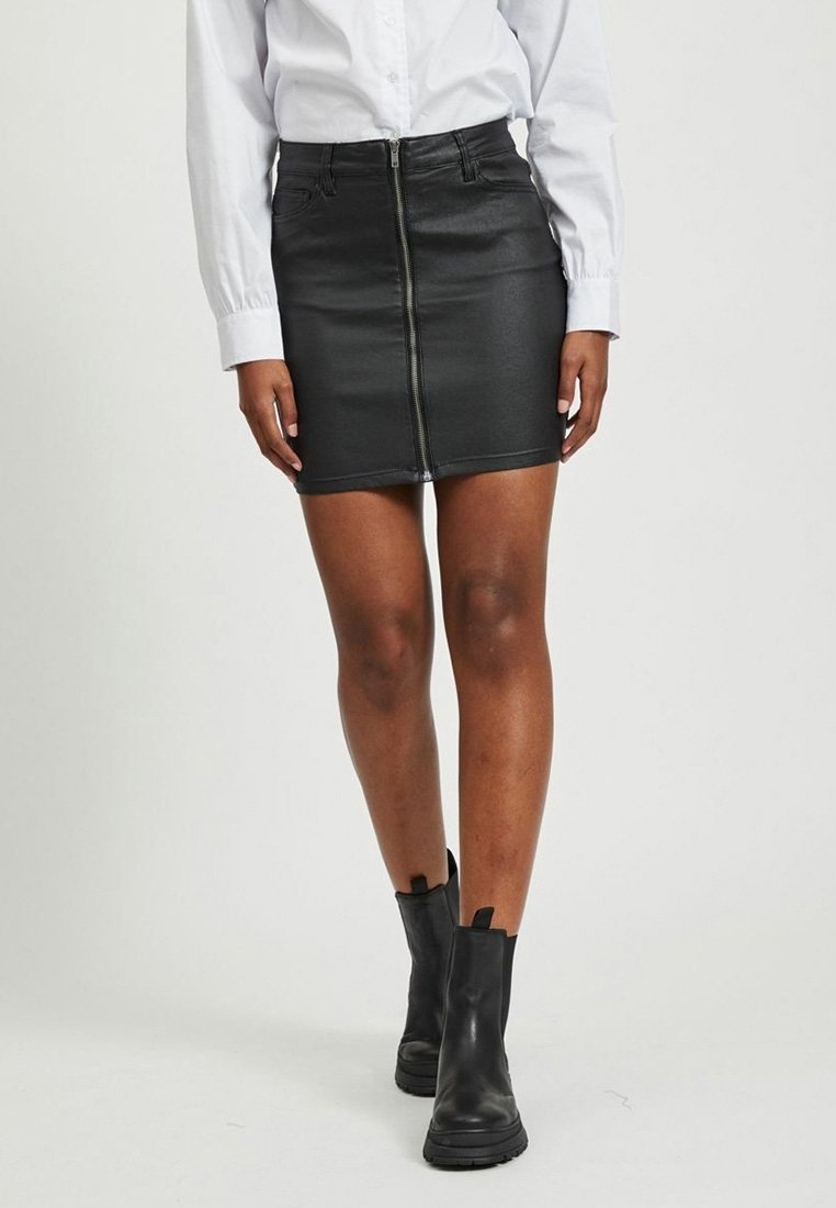 Femme portant une mini-jupe en similicuir noire avec fermeture éclair devant, chemise blanche à manches longues et bottines noires, debout devant un fond uni.