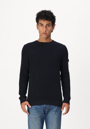 Navyblauer Strickpullover mit geripptem Muster, Rundhalsausschnitt und langen Ärmeln, der ein kleines Logo auf dem linken Ärmel trägt. Kombiniert mit blauen Jeans.