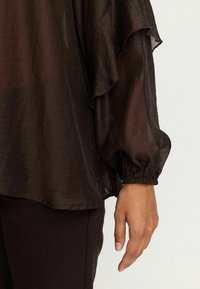 Blouse marron, transparente avec des volants superposés et des manches volumineuses élastiquées. Portée avec un pantalon sombre assorti. Texture lisse.