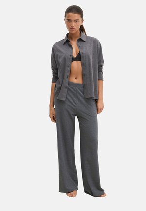 Vrouw staat blootsvoets, draagt losse grijze broek, een open grijs overhemd met knopen en een zwarte bralette, tegen een effen witte achtergrond.