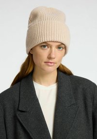 Lichtbeige geribbelde gebreide beanie met een omgeslagen rand, zachte textuur, gedragen met een donkergrijze jas over een wit shirt.