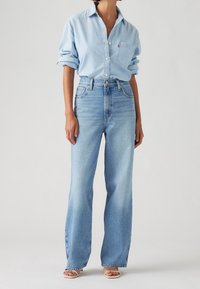 Vaaleansininen farkkupaita, jossa on napitettava etuosa, pitkät hihat ja rintatasku, yhdistettynä korkeavyötäröisiin vaaleansinisiin leveälahkeisiin farkkuihin.