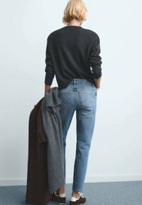 Maglione lavorato a maglia scuro, jeans a vita alta azzurri chiari e cappotto scuro appoggiato sul braccio, con scarpe marroni con dettagli bianchi.