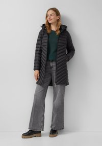 Zwarte gewatteerde pufferjas met een capuchon, voorzien van een rits en diagonale stiksels. Draagt over een groene trui en grijze wijde jeans.