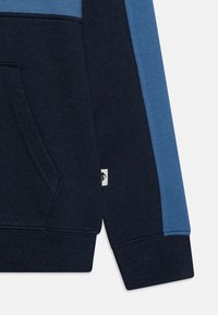 Sudadera con capucha en azul marino y azul claro, con un bolsillo y puños acanalados, hecha de un tejido texturizado y con una etiqueta de logo distintiva.