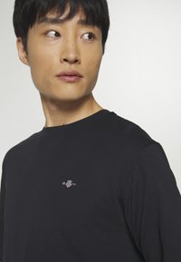 Camiseta negra de manga larga hecha de tela suave, con un pequeño logotipo bordado en el pecho. Cuello redondo con un ajuste holgado.