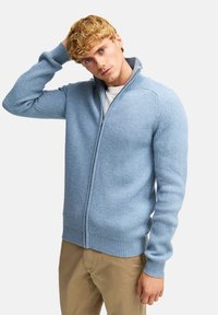 Jasnoniebieski prążkowany sweter zapinany na zamek z wysokim kołnierzem i dopasowanym krojem. Rękawy i dół mają prążkowane mankiety, wykonane z miękkiego przędzy.