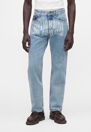 Lyseblå jeans med falmet hvid "Billionaire Boys Club" tekst båret af en person, der står, kombineret med mørkebrun lædersko.