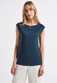 Top senza maniche color teal con scollatura rotonda e dettaglio a chiave, realizzato in tessuto morbido e con una vestibilità morbida, abbinato a pantaloni chiari.