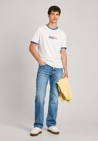 Balta trumpa rankovė t-shirt marškinėliai su mėlynais akcentais ir grafiniais spausdinimais, derinami su šviesiai mėlynomis džinsomis ir baltais batais, laiko geltoną megztinį.