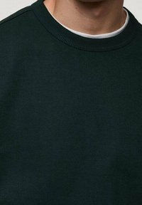 Mężczyzna w ciemnozielonym swetrze z okrągłym dekoltem nosi pod nim białą koszulę z okrągłym dekoltem, odsłaniając szyję i górną część klatki piersiowej.