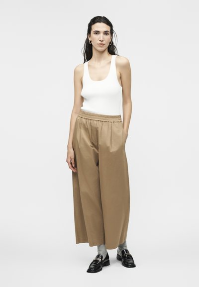 Γυναίκα με μακριά σκουρόχρωμα μαλλιά που φοράει λευκό αμάνικο τοπ, φαρδιά μπεζ παντελόνια, γκρι κάλτσες και μαύρα μοκασίνια, μπροστά από μια μονόχρωμη φόντο.