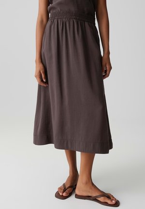 Frau trägt einen dunkelbraunen Midirock mit elastischem Bund und passenden braunen flachen Sandalen, steht auf einem hellgrauen Boden.