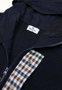 Giacca con zip blu navy con trama a coste e una toppa in tessuto a quadri. Presenta un cappuccio e un'etichetta con il marchio "Aqua Scutum".