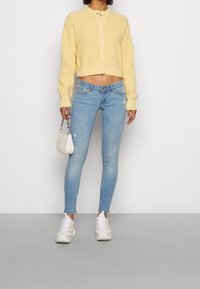 Gul kortärmad stickad cardigan med rund hals och knappstängning, i kombination med ljusblå skinny jeans och vita sneakers. Liten vit handväska.