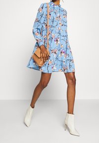 Kvinna klädd i en blå blommig klänning, vita ankelstövlar och bär en beige axelväska, står framför en enkel vit bakgrund.