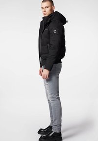 Czarna kurtka puchowa z kapturem, zapinana na zamek, z prążkowanymi mankietami i naszywką z logo. Noszona z jasnoszarymi dżinsami z przetarciami i czarnymi masywnymi butami.