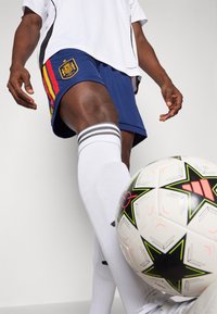 Μπλε ποδοσφαιρικές βερμούδες με το εθνικό έμβλημα της Ισπανίας, με κόκκινες και κίτρινες ρίγες. Λευκές κάλτσες με μαύρες ρίγες δίπλα σε μια σχεδιασμένη μπάλα ποδοσφαίρου.