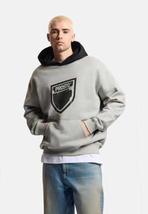 Młody mężczyzna z krótkimi blond włosami, ubrany w szarą bluzę Prosto na czarnej bluzie z kapturem i niebieskich dżinsach, ręce w kieszeni bluzy.