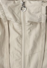 Primer plano del frente de una chaqueta beige con cremallera central y cordones de tela a cada lado.