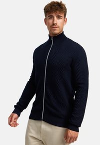 Marineblå zip-up genser med teksturert sikksakkmønster, ribbestrikkede mansjetter og kant, samt høy krage. Laget av et mykt, strikket materiale.