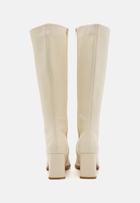 Bottes montantes crème en matériau synthétique lisse, avec un talon carré et une fermeture éclair arrière pour un enfilage facile. Design minimaliste.