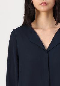 Blouse bleu marine foncé avec une texture lisse, dotée d'un design sans col et d'un détail subtil de couture frontale. Le tissu a un léger éclat.