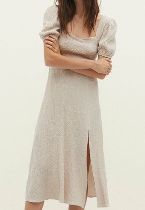 Vrouw draagt een beige midi-jurk met textuur, pofmouwen en een zijsplit, staand met gekruiste armen tegen een effen achtergrond.