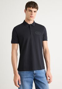 Camisa polo preta, feita de algodão suave, com colarinho, mangas curtas e um logótipo bordado no peito. Usada com calças de ganga azuis.