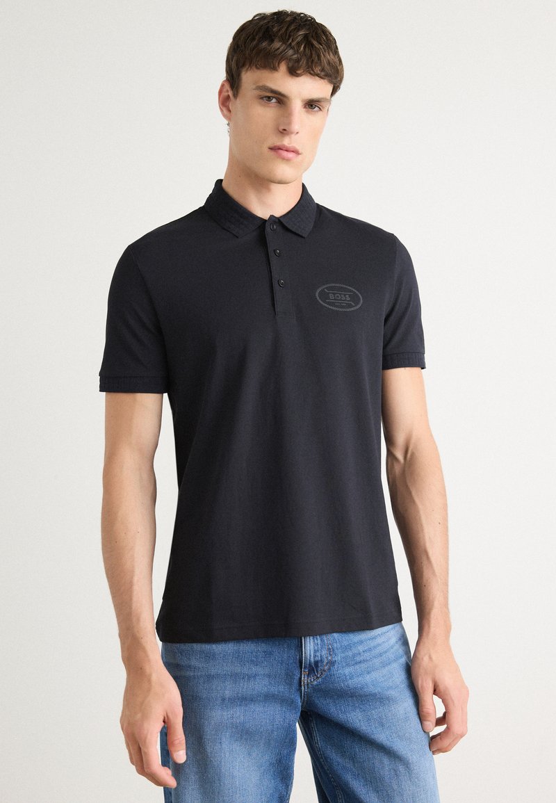 Camisa polo preta, feita de algodão suave, com colarinho, mangas curtas e um logótipo bordado no peito. Usada com calças de ganga azuis.