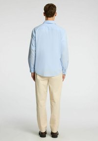 Camisa de textura azul claro con mangas largas y puños abotonados, combinada con pantalones color crema y zapatos marrones. Diseño limpio y casual.
