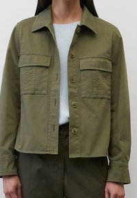 Persona que lleva una chaqueta abotonada verde oliva con bolsillos de solapa sobre un suéter de punto gris claro y pantalones verdes oliva a juego.