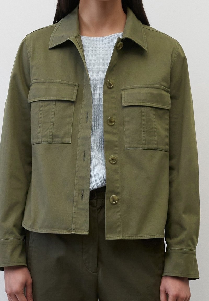 Persona que lleva una chaqueta abotonada verde oliva con bolsillos de solapa sobre un suéter de punto gris claro y pantalones verdes oliva a juego.