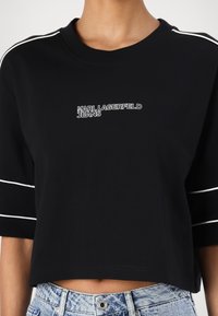 Melnā īsā teļa kreklā ar baltām sānu svītrām, kas rotāta ar tekstu "KARL LAGERFELD JEANS". Kokvilnas materiāls ar gludu tekstūru.