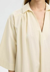 Camisa beige de gran tamaño con cuello doblado, mangas cortas y sutiles pliegues. Fabricada en suave y ligero algodón.
