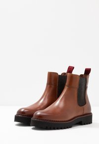 Une paire de bottes Chelsea en cuir marron avec des panneaux latéraux en élastique noir, des semelles noires épaisses et des languettes rouges à l'arrière sur fond blanc.