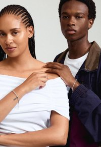 Dos personas están vestidas de manera casual. La mujer tiene el cabello trenzado y lleva una blusa blanca sin hombros, mientras que el hombre usa una chaqueta oscura y una camisa blanca.