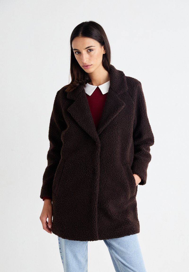 Un manteau brun, duveteux, à texture avec un col cranté et des poches latérales, superposé à un haut rouge avec un col blanc.