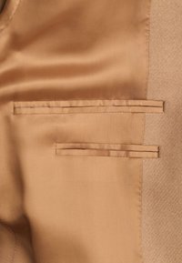 Intérieur d'une veste couleur chameau montrant une doublure en satin lisse avec deux poches horizontales passepoilées sur un panneau de tissu texturé.