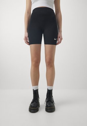 Osoba ubrana w czarne spodenki rowerowe Nike, biały prążkowany krótki top, białe skarpetki i czarne sznurowane buty, stojąca na jednolitym jasnym tle.