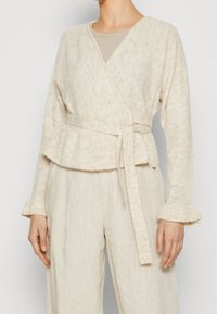 Wrap sweater in a soft, cream textured knit with a peplum hem and tie belt; paired with loose, light-colored linen pants.