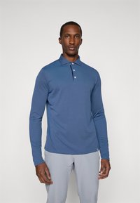 Polo majica z dolgimi rokavi v modri tkanini z ovratnikom, tremi gumbi in ožjim krojem. Kombinirana s svetlo sivo hlačami, ima gladko teksturo.