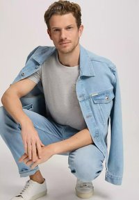 Jasnoniebieska dżinsowa kurtka na szarej koszuli, zestawiona z jasnoniebieskimi dżinsami i białymi trampkami. Kurtka ma zapięcie na guziki z przodu oraz kieszenie.