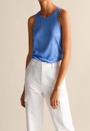 Ärmelloses blaues Tanktop kombiniert mit weißer, hoch taillierter Hose mit seitlichen Taschen an einer stehenden Person.