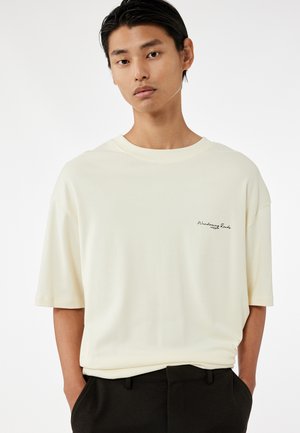 Cremfarbener, kurzärmeliger T-Shirt aus einem weichen Stoff, mit einem kleinen schwarzen gestickten Logo auf der linken Seite. Gerade geschnittenes Design.