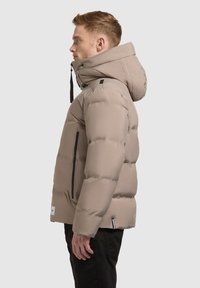 Beige pufferjack met een hoge kraag en grote capuchon. Voorzien van ritssluitingen, gestikte panelen en een licht gewatteerde textuur.