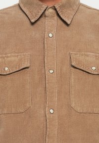 Bruine corduroy shirt met een knoopsluiting, zachte textuur, kraag en twee borstzakken. Voorzien van zilveren drukknoopjes en verticale ribbels.