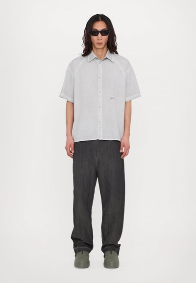Chemise à manches courtes gris clair avec un logo subtil, associée à un jean large gris foncé et des chaussures vertes. Design minimaliste et coupe décontractée.